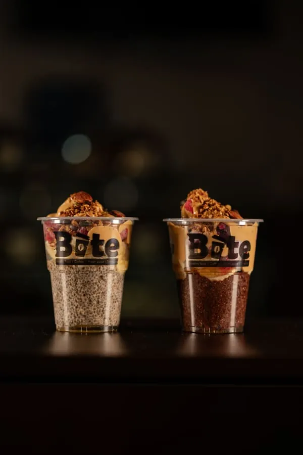 Two BITE chia pudding cups side by side, one plain and one with cocoa, both topped with granola, peanut butter, almonds and dried berries.