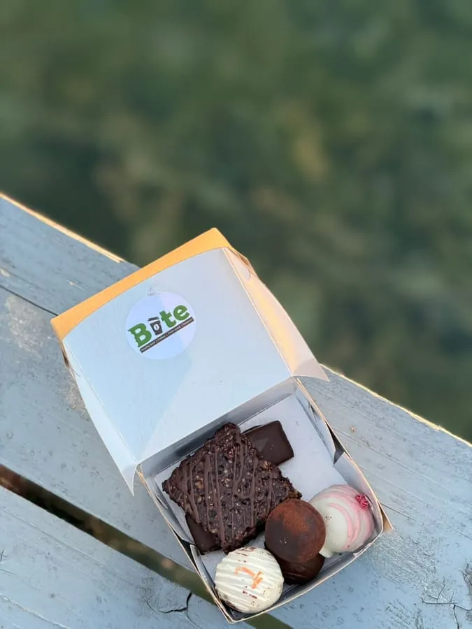 An open BITE box of chocolate truffles and a brownie, balanced on a white wooden railing above Lake Ohrid.