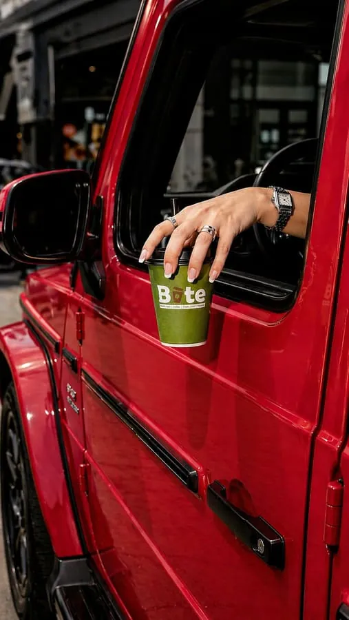 A hand reaching out of a red car window to take a green BITE takeaway cup resting on the door.